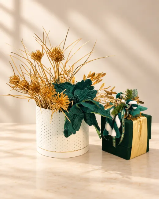 Scatolina regalo in velluto verde con nastro oro, accanto a un vaso bianco con rami e fiori dorati
