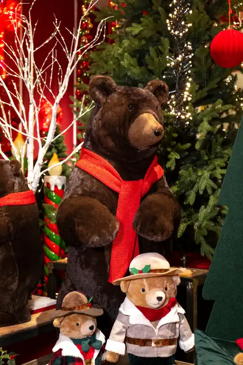 Un grande orso di peluche con la sciarpa rossa e due orsetti vestiti di tartan, fra abeti, rami bianchi e sfere rosse